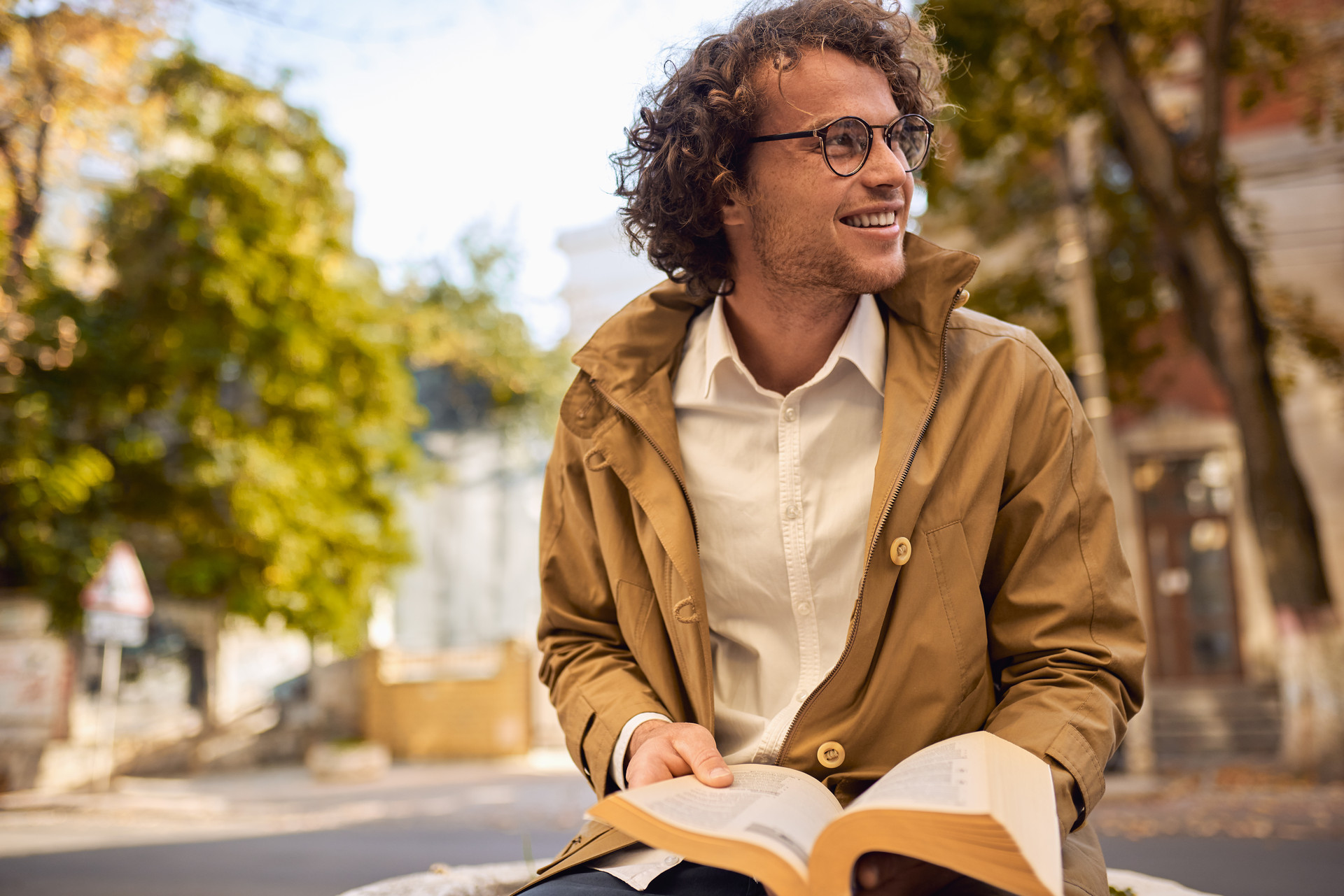 Ein Mann sitzt auf einer Mauer und liest ein Buch. Er trägt eine braune Jacke und eine Brille.